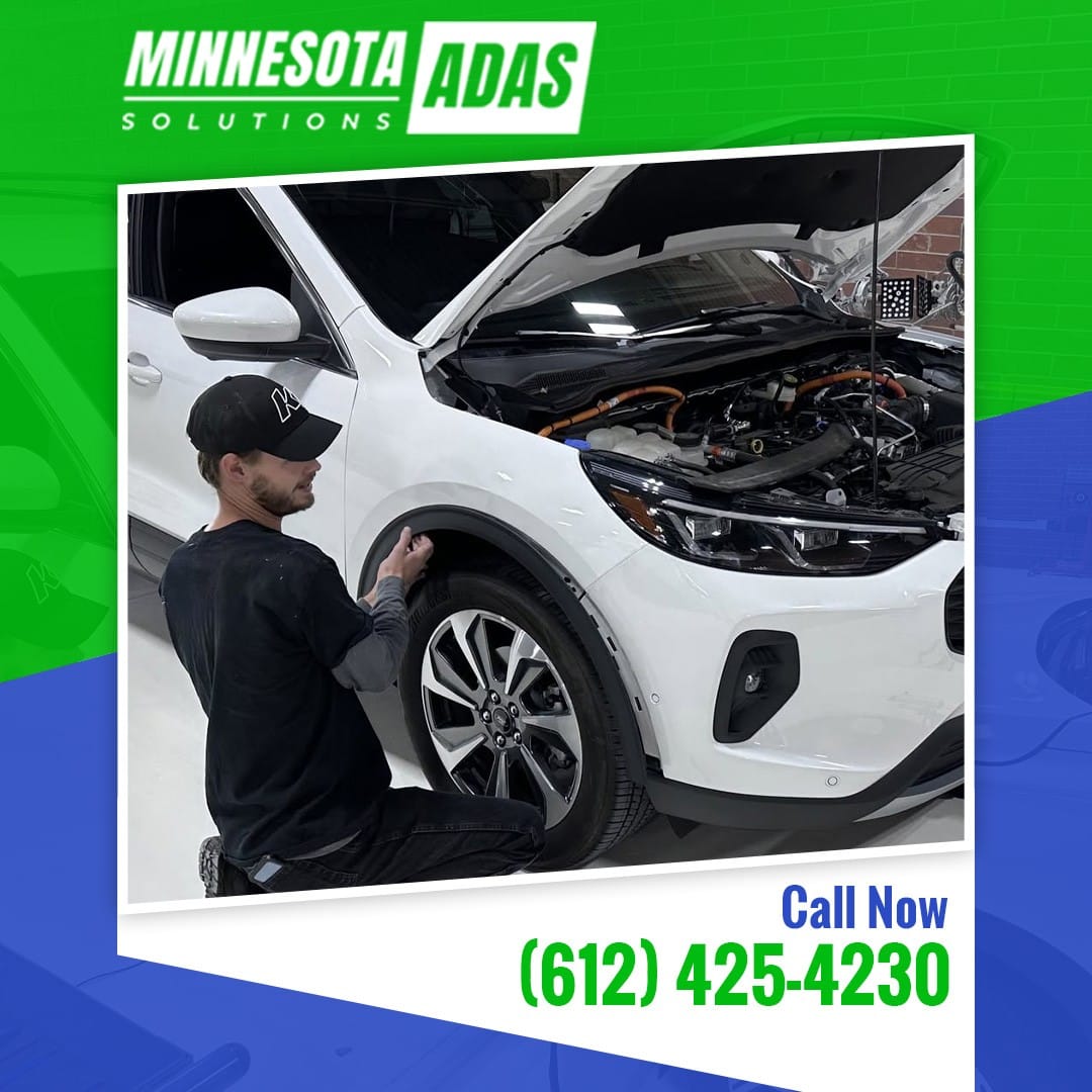 Automotive technician kneeling near the open hood and front wheel well of a white car during sensor diagnostics.